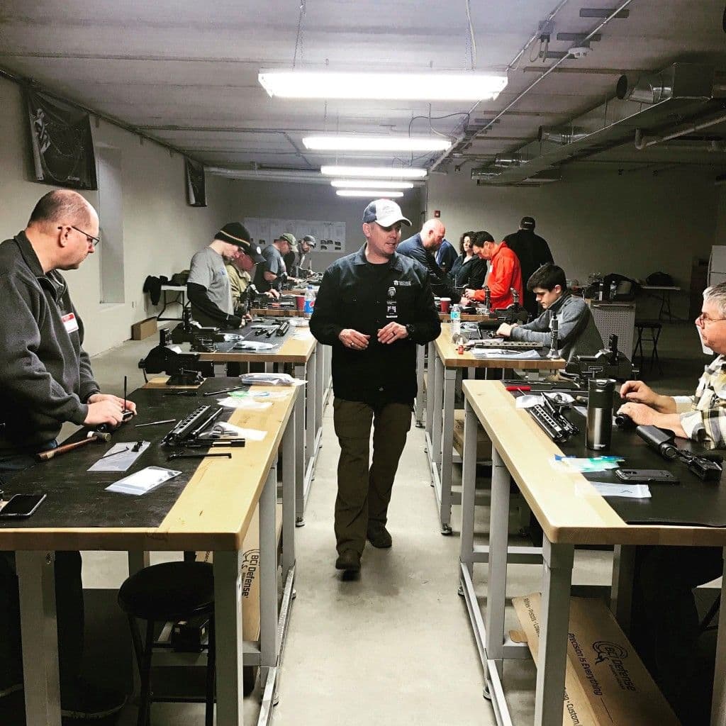 Students working at parallel armorer benches in a training workshop while an instructor walks the center aisle.