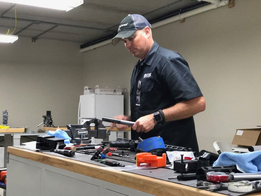 An armorer examining a barrel at a well-equipped workbench with vises and hand tools.